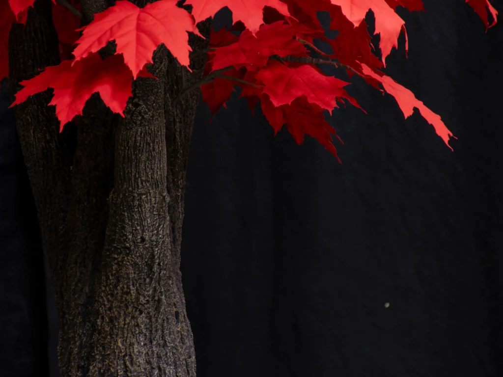 Artificial maple tree trunk with rough dark bark and bright red autumn leaves extending from branches.